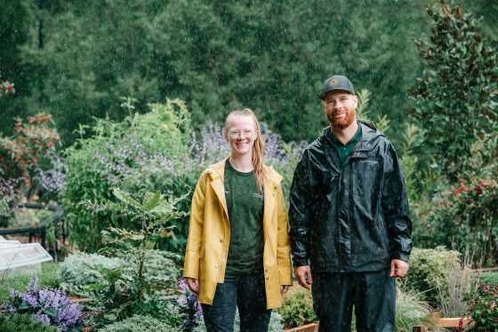Two people standing shoulder to shoulder, in the rain, in front of an outdoor model train display. 